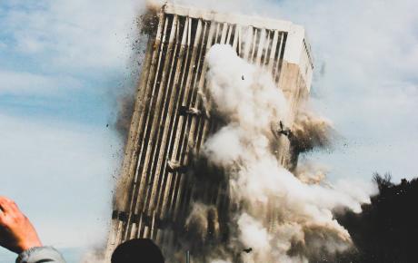 An office building collapses in the background while, in the foreground, several people take photos of the catastrophe on their phone