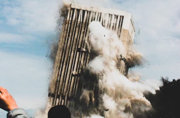 An office building collapses in the background while, in the foreground, several people take photos of the catastrophe on their phone